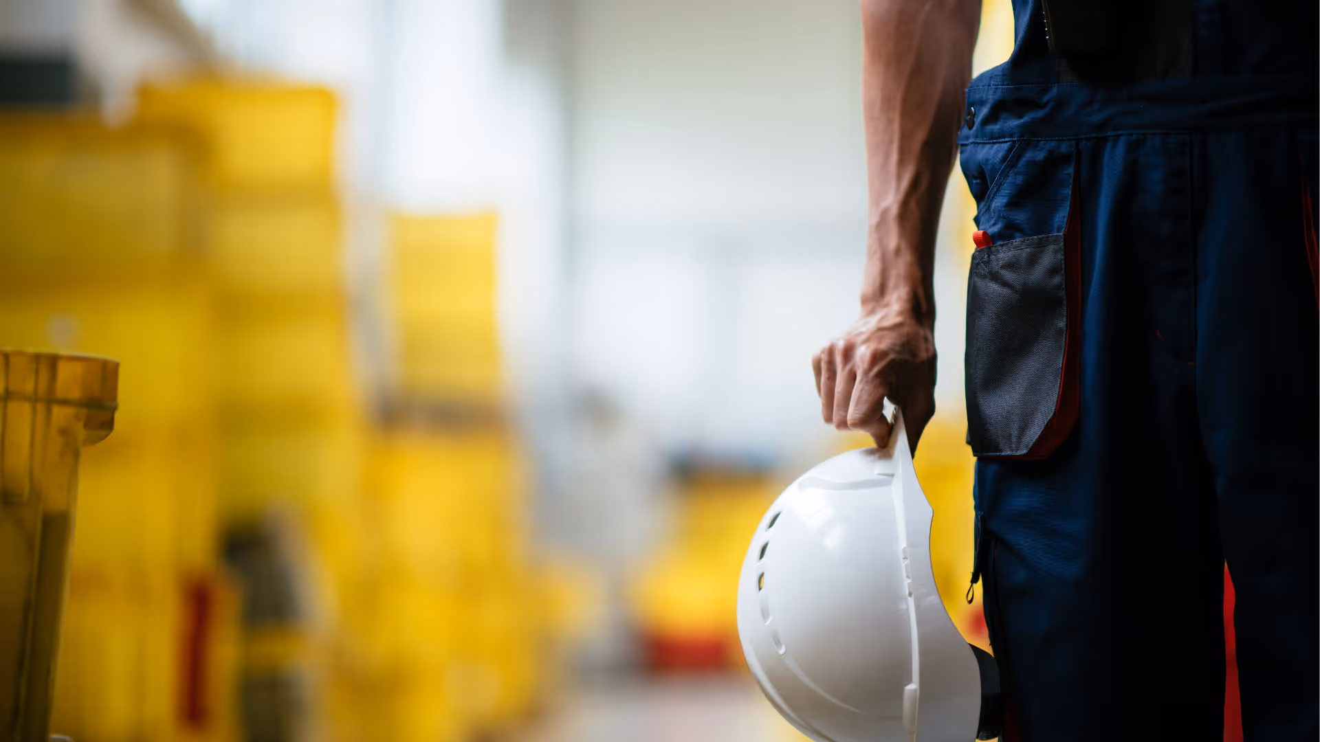 Person in dark work overalls holding a white safety helmet with blurred yellow containers in the background.
