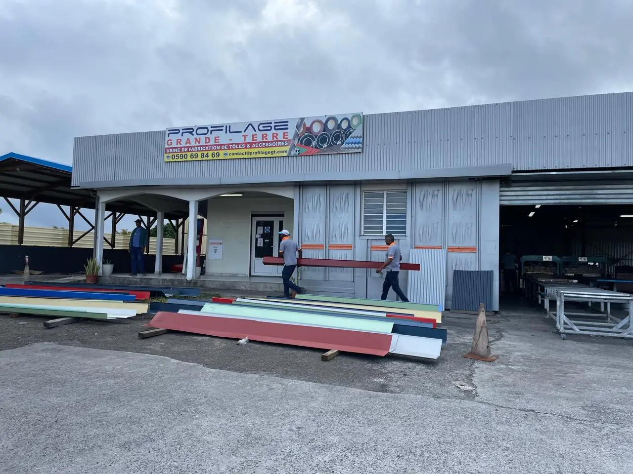 Deux hommes portant une poutre devant une usine de fabrication de tôles et accessoires avec plusieurs tôles colorées posées au sol.