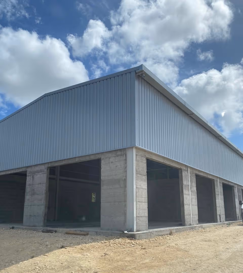 Bâtiment industriel en construction avec une structure en béton et un toit en métal sous un ciel partiellement nuageux.