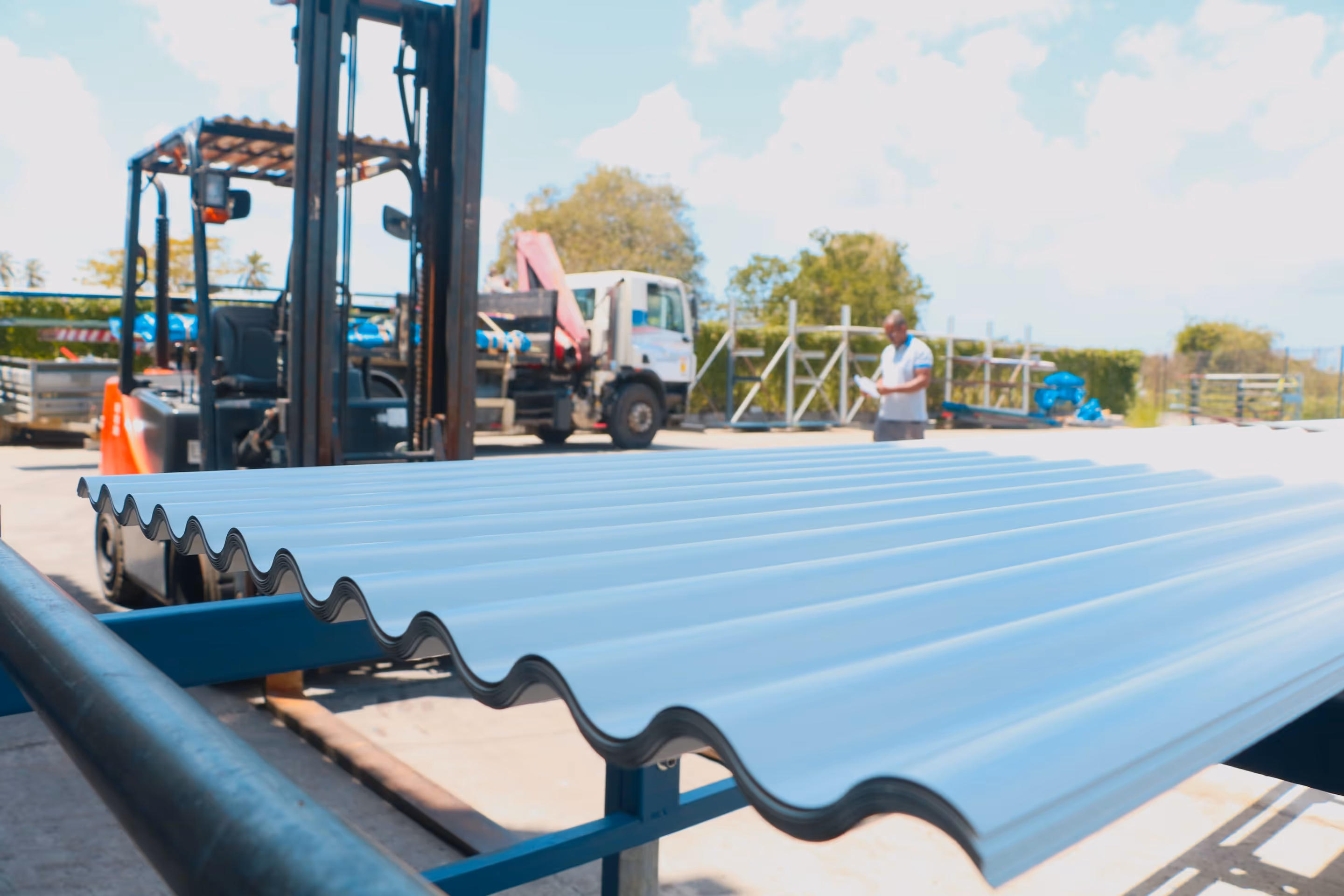 Plaques ondulées blanches en métal posées sur une structure métallique dans un chantier extérieur avec un chariot élévateur et un ouvrier en arrière-plan.