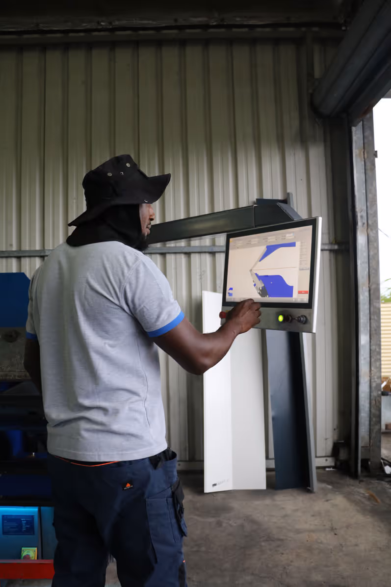 Un homme en chapeau noir utilise une machine à commande numérique avec un écran affichant un logiciel de modélisation 3D dans un atelier industriel.