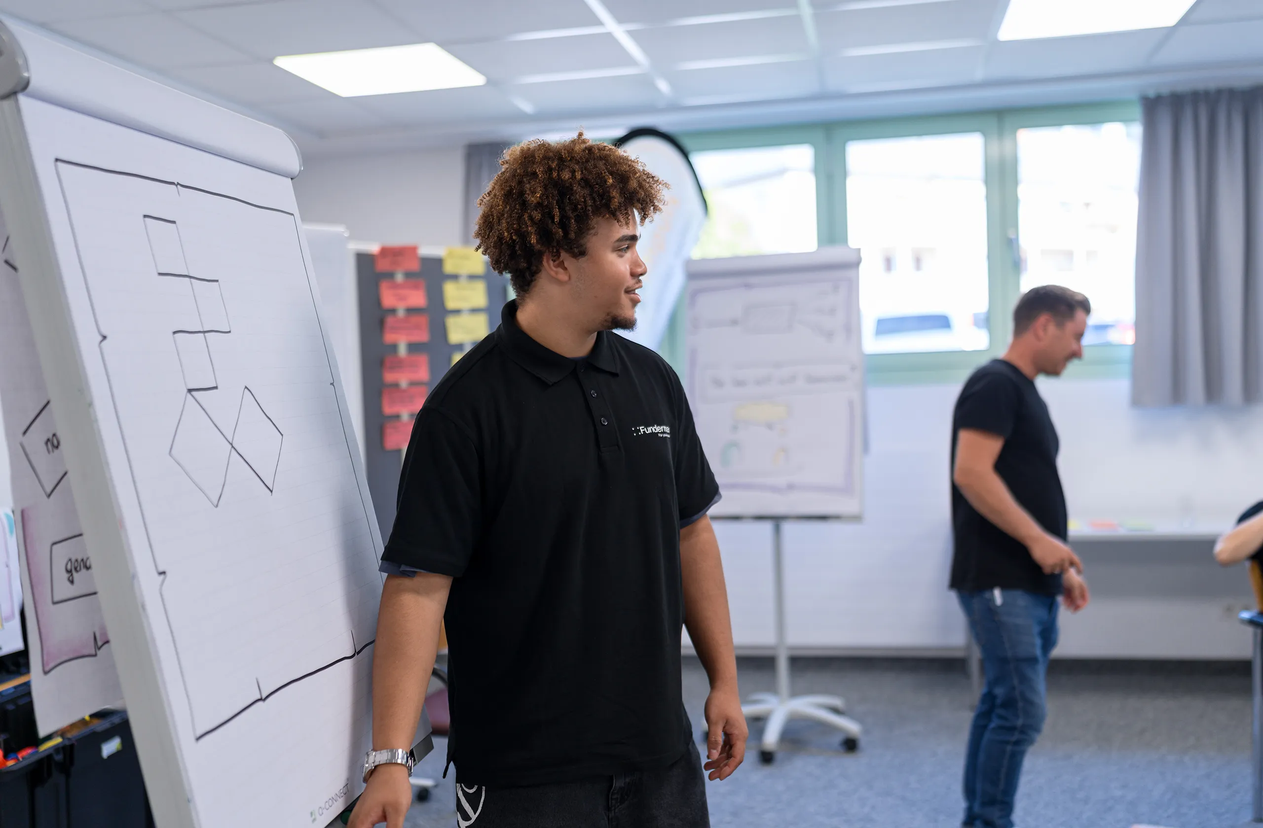 Lehrling von Fundermax mit schwarzem Polo präsentiert lächelnd am Flipchart mit Simon Schelkshorn im Hintergrund erkennbar.