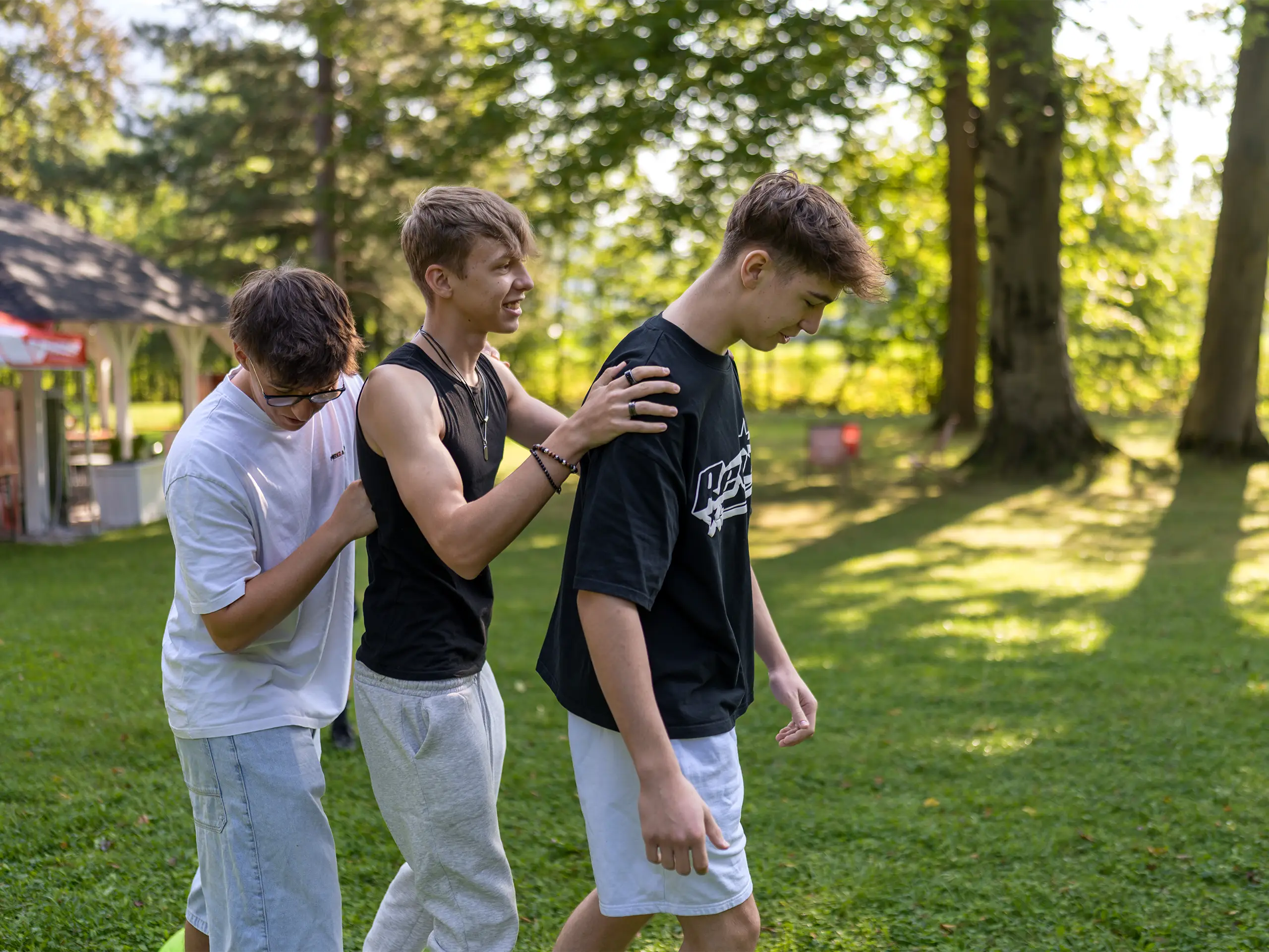 Drei männliche Lehrlinge absolvieren bei einem Lehrlingscamp im Park eine Teambuilding-Übung und lachen gemeinsam.