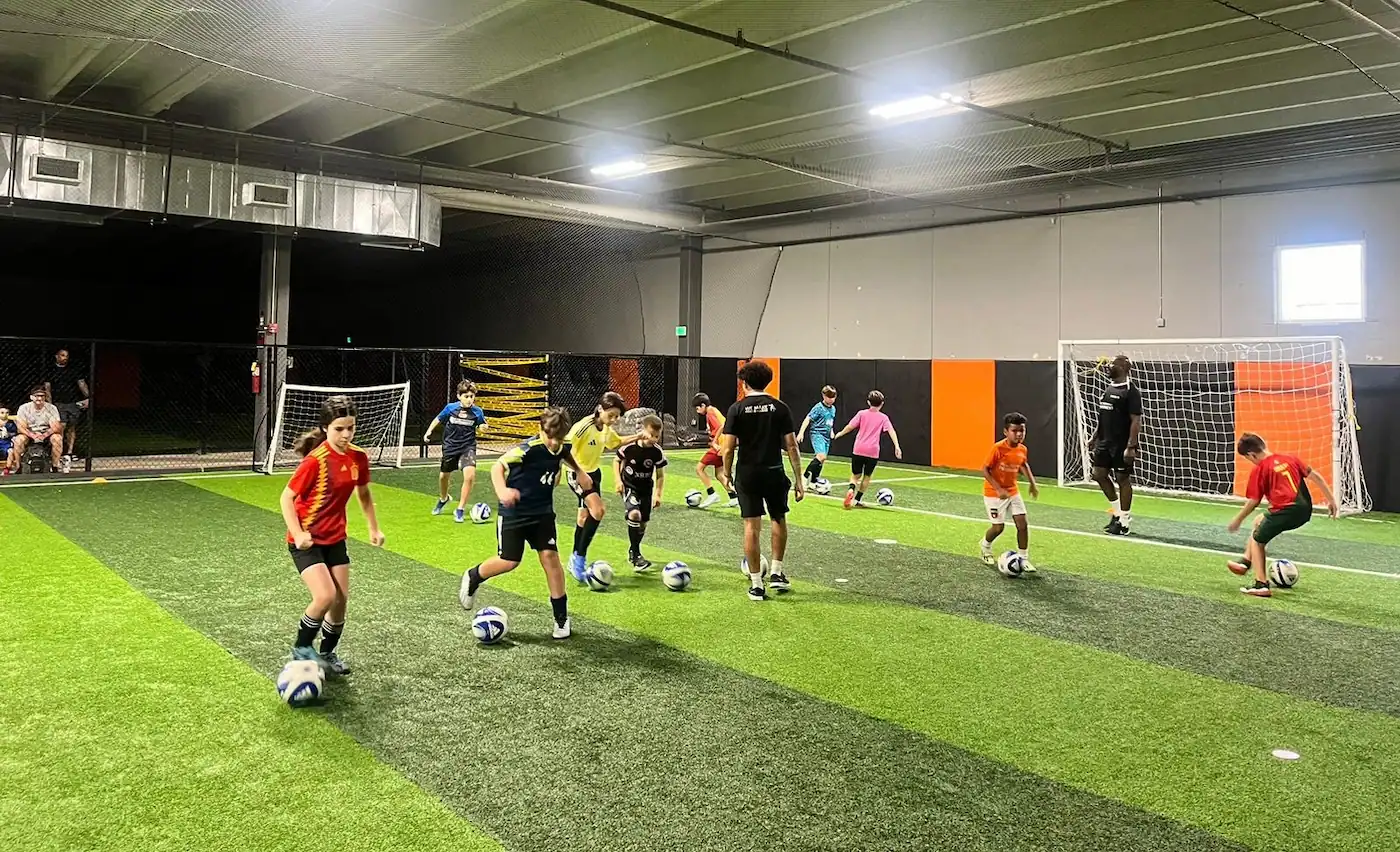 Children playing a structured soccer training session on an indoor pitch, led by a coach wearing black training gear, with mini goals and soccer balls spread across the pitch.