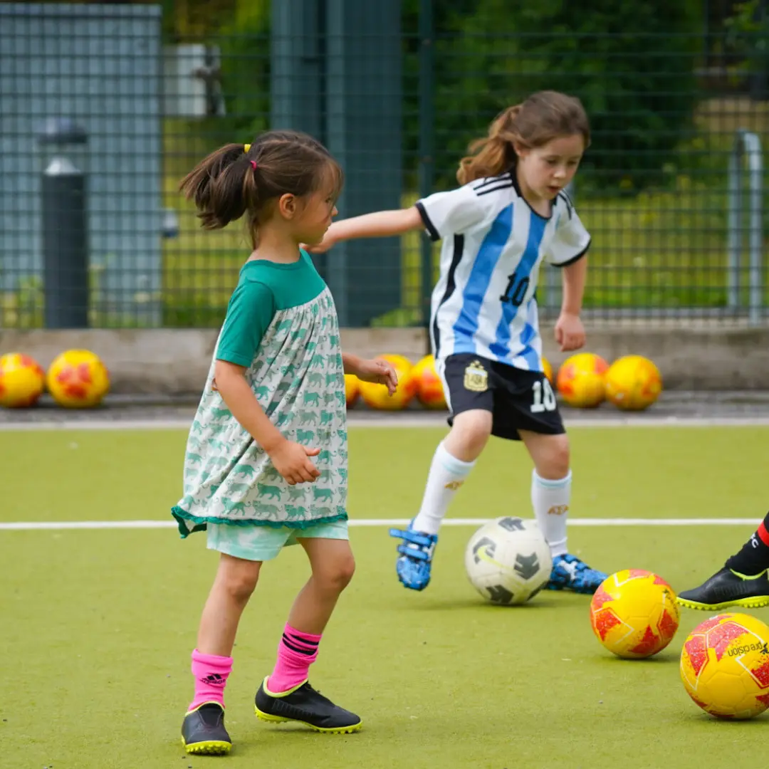 Two girls practising football drills with several balls on an outdoor pitch
