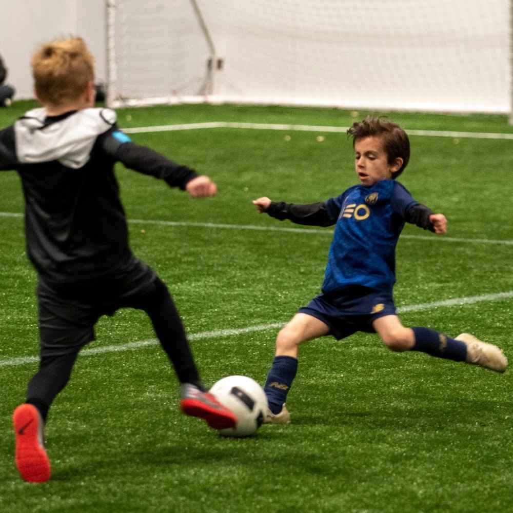 Two boys sliding to tackle a football on an indoor pitch
