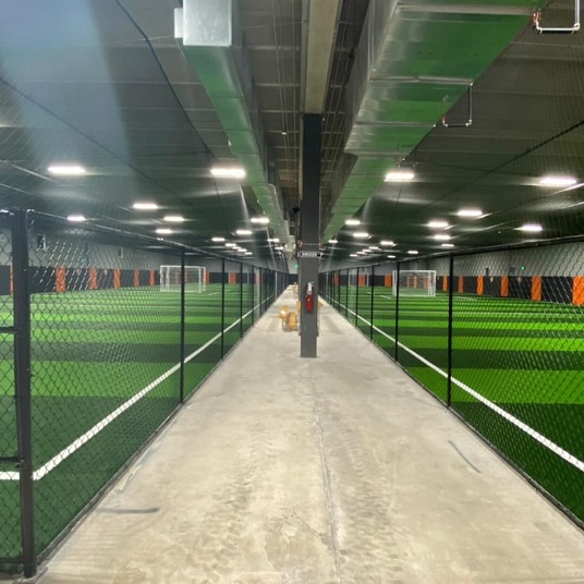 Indoor corridor between fenced football pitches lit by overhead lights
