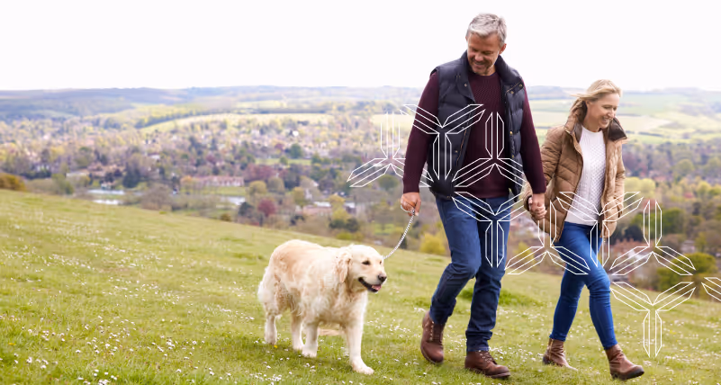 A middle aged couple are walking their dog outside in a field in cool weather.