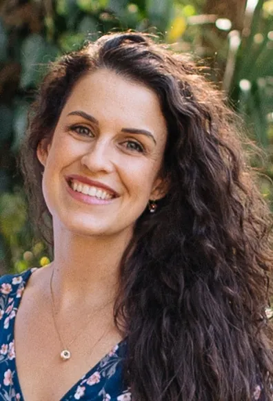 Smiling woman with long curly dark hair wearing a blue floral dress and a necklace, standing outdoors with greenery in the background.
