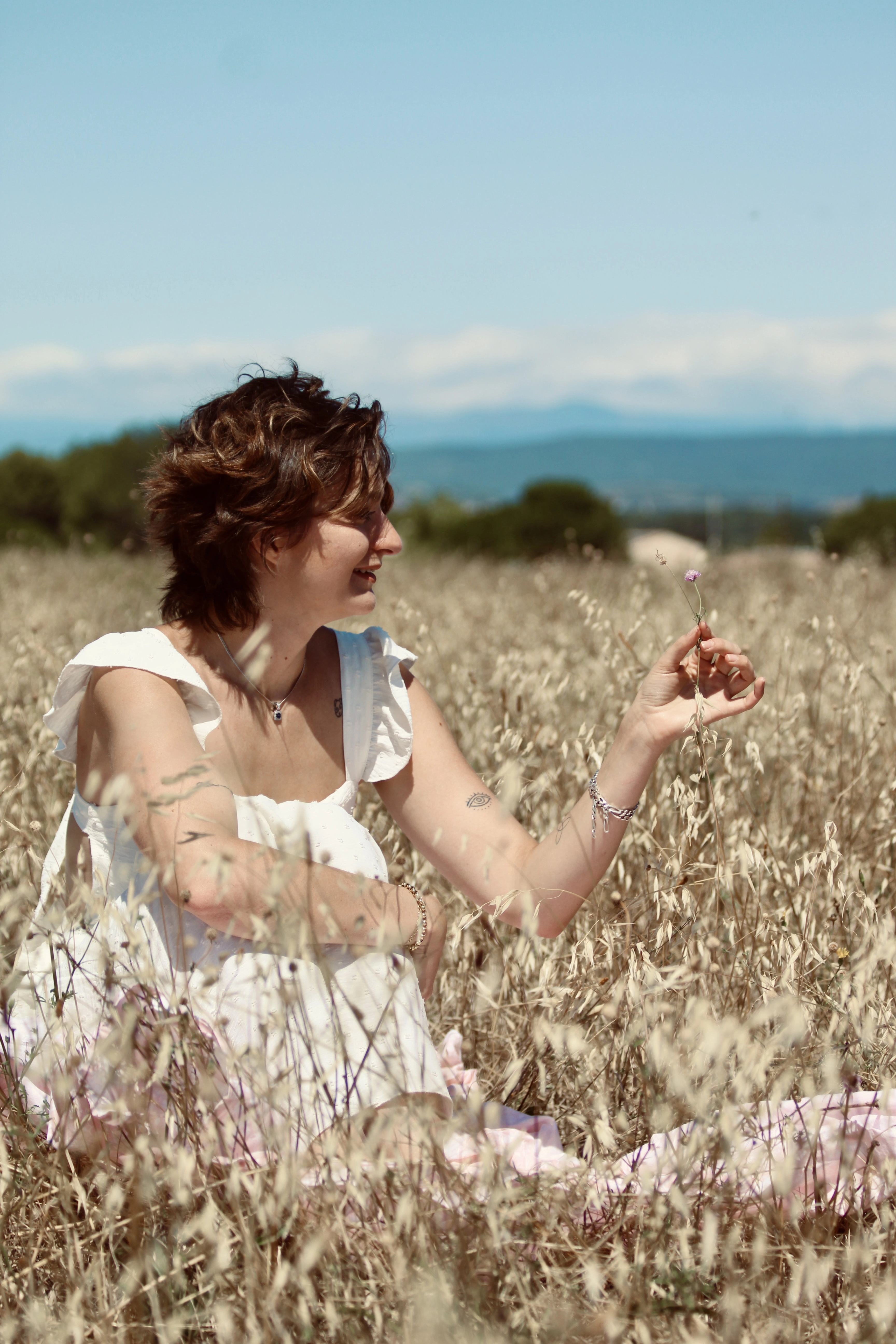 Femme assise dans un champ d’herbes sèches, souriant et observant une petite fleur entre ses doigts.