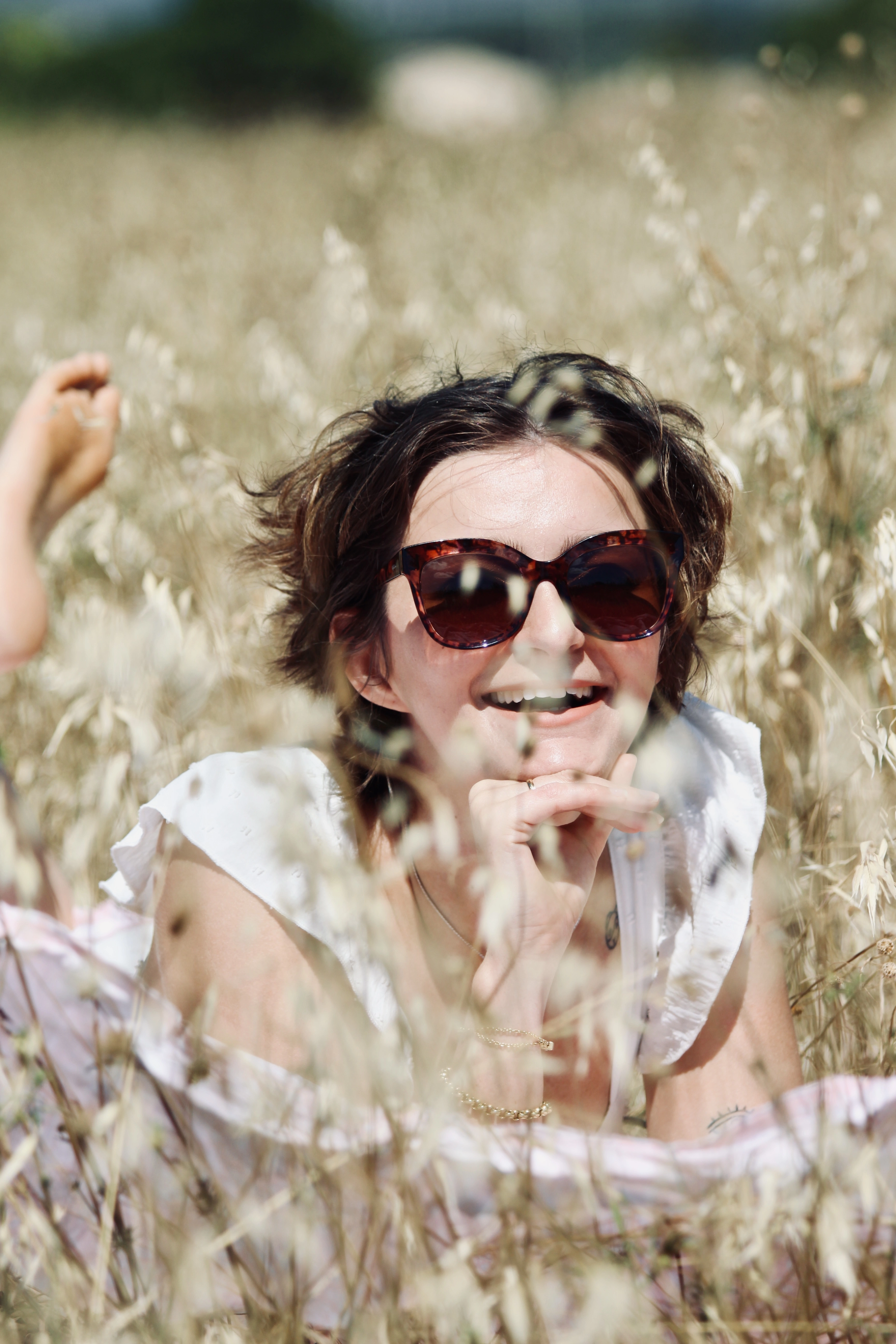 Femme allongée dans un champ, souriante, portant des lunettes de soleil, entourée d’herbes hautes.