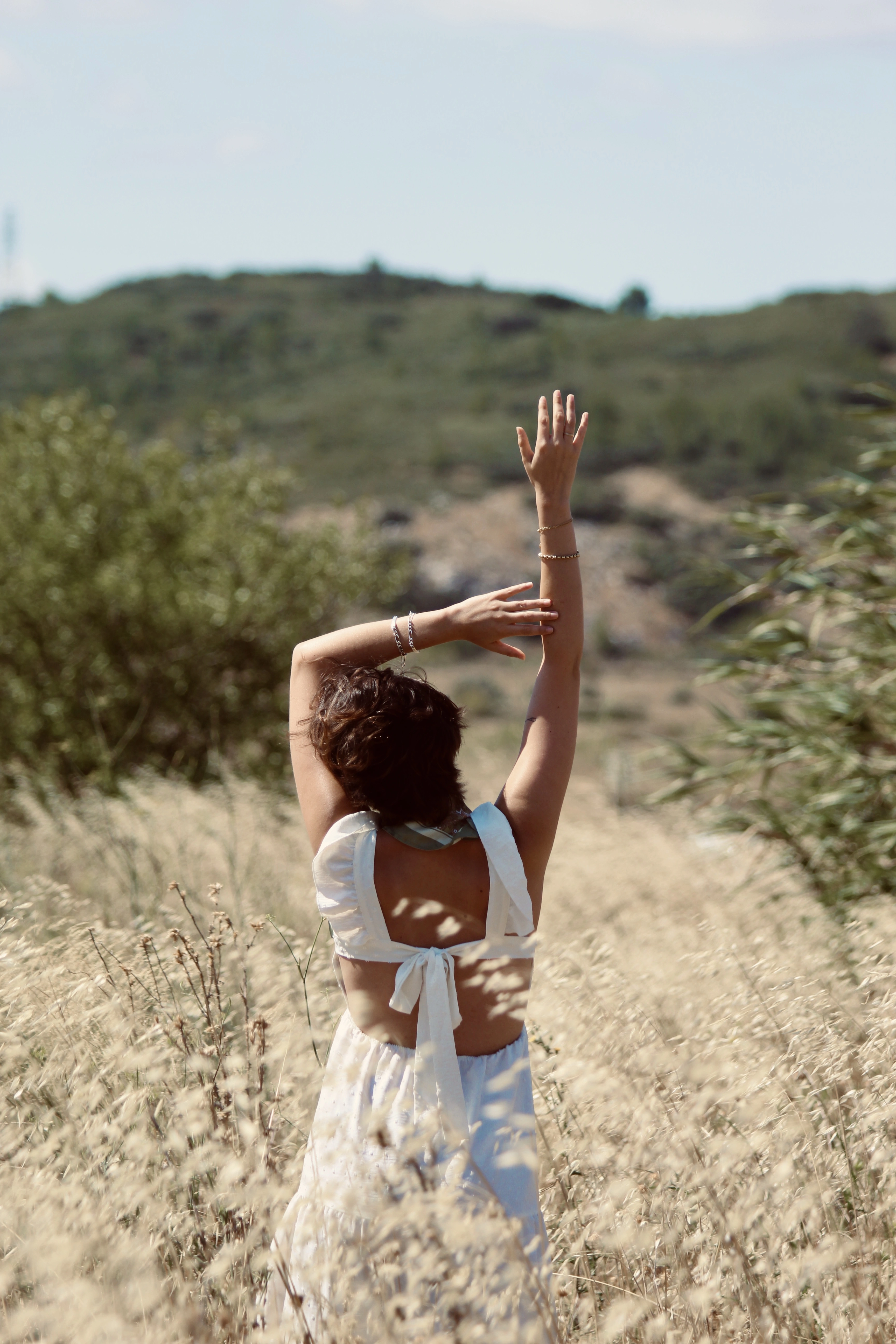 Femme en robe blanche levant les bras vers le ciel en marchant dans un champ, vue de dos.