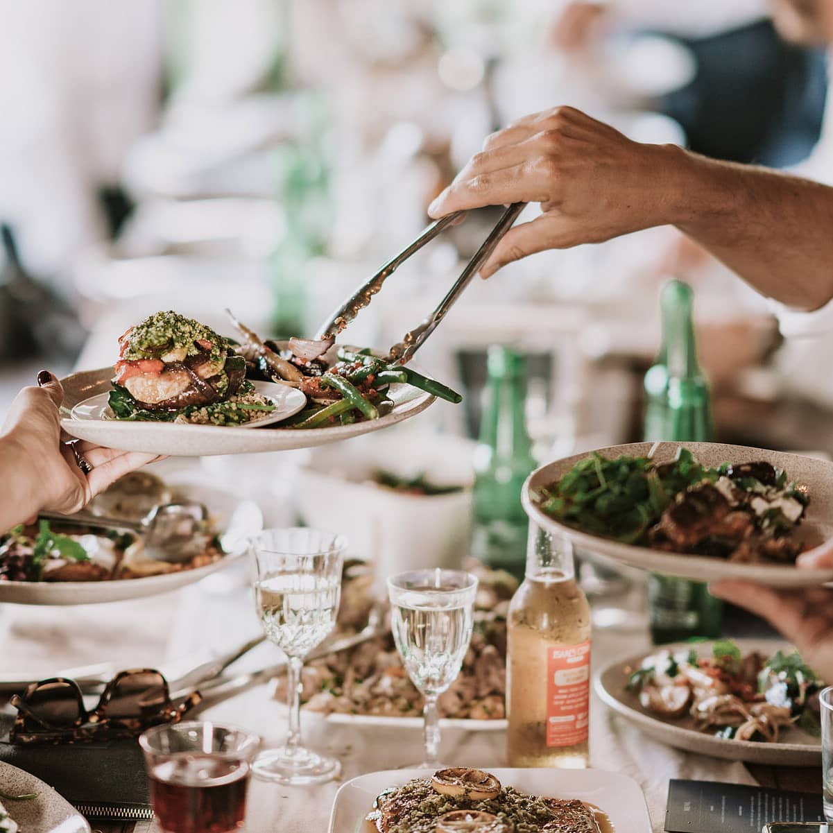 A photo of someone using tongs placing some vegetables onto someones plate. The table is full of delicious looking sharing food.
