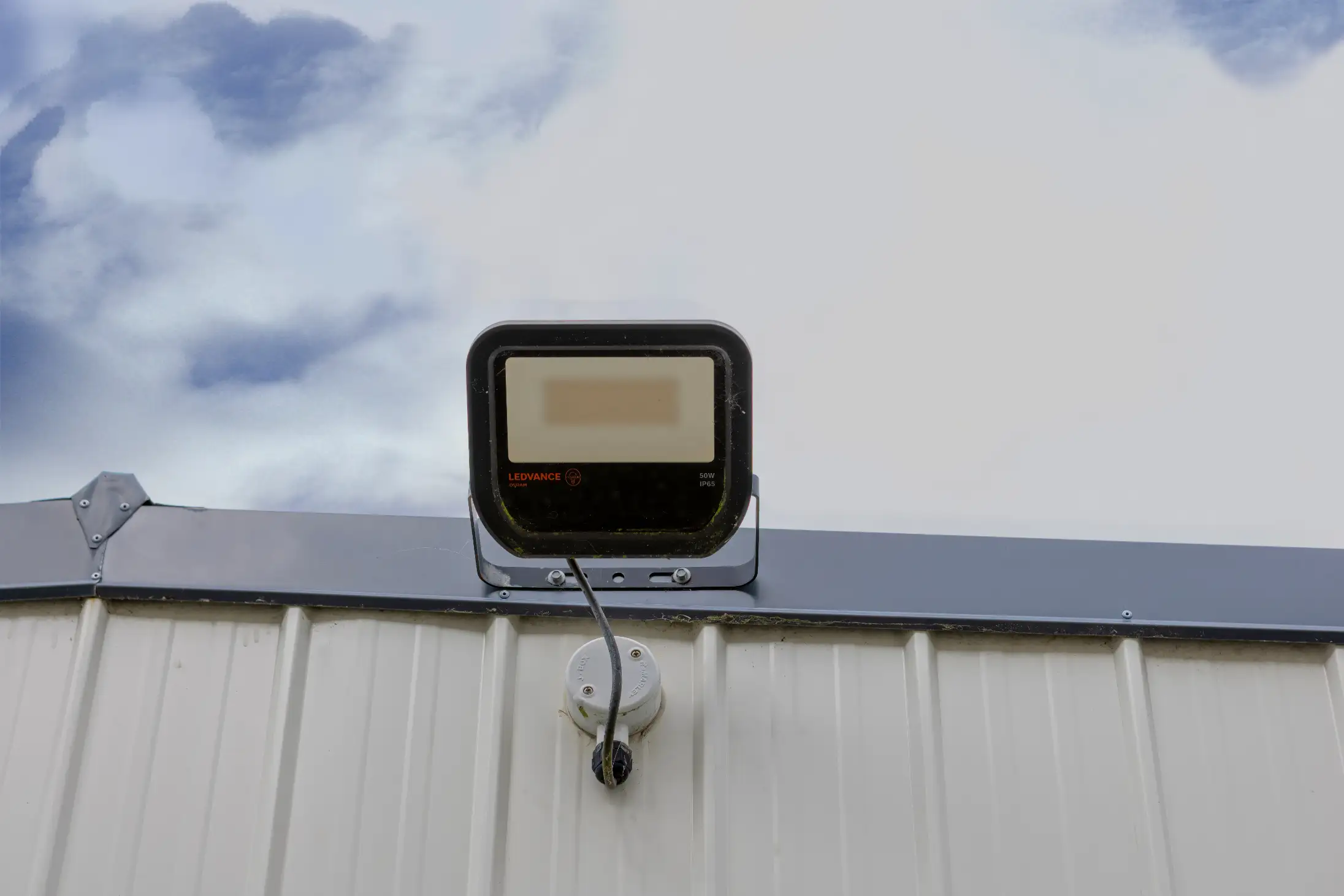 A rectangular LED floodlight is mounted on the exterior wall of a metal building, against a cloudy sky. A black power cord connects the light to a small junction box below it.
