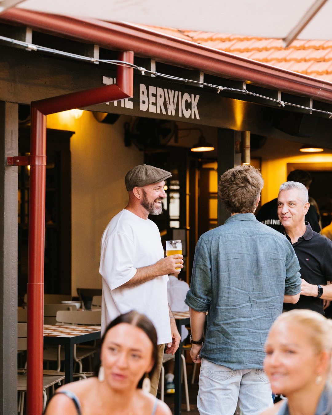 Three men socializing outside a pub called The Berwick, one holding a glass of beer and smiling, with two women slightly blurred in the foreground.