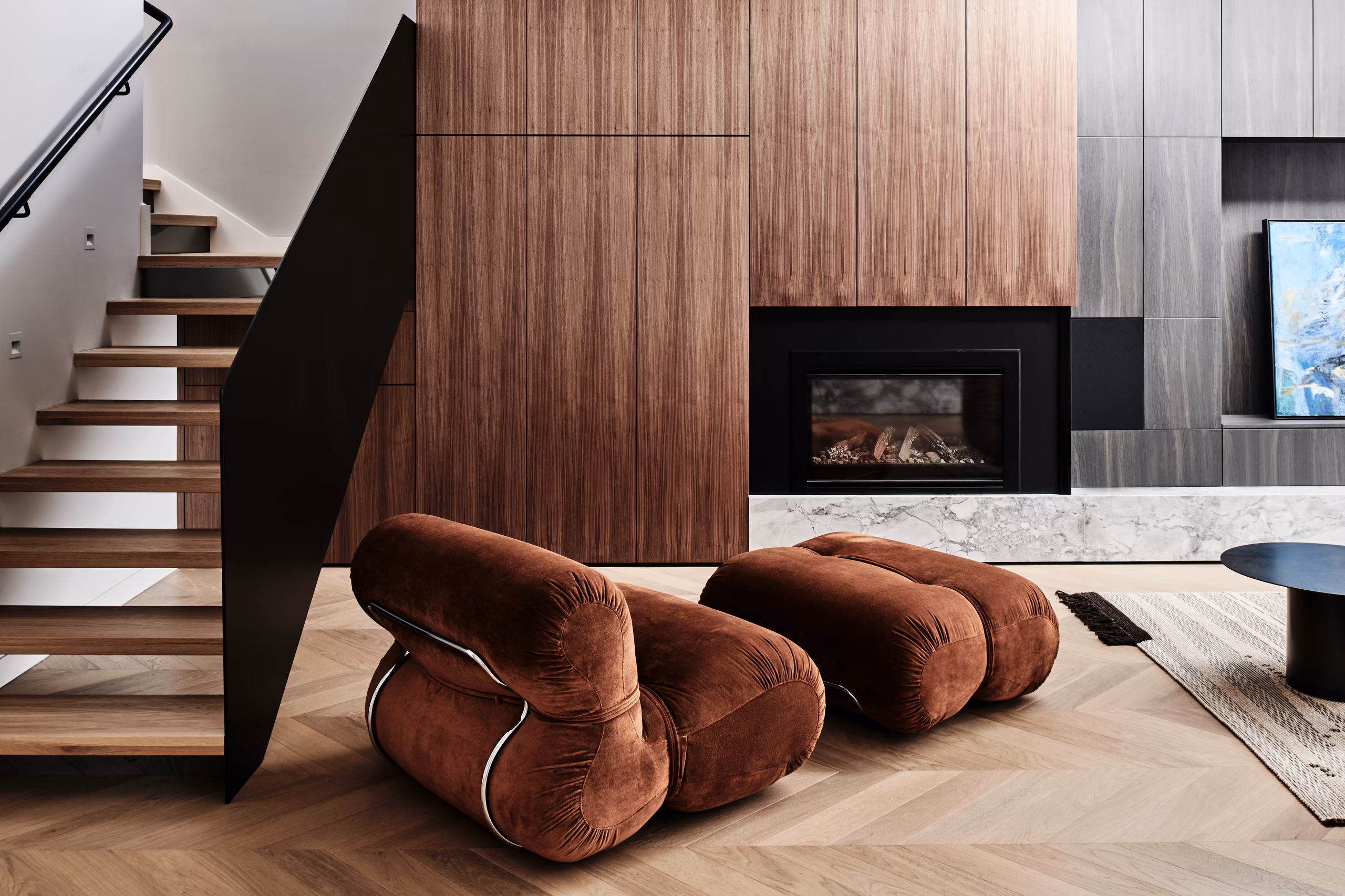 living room with rust velvet lounge chair and ottoman, walnut-toned joinery, fireplace and open timber stair