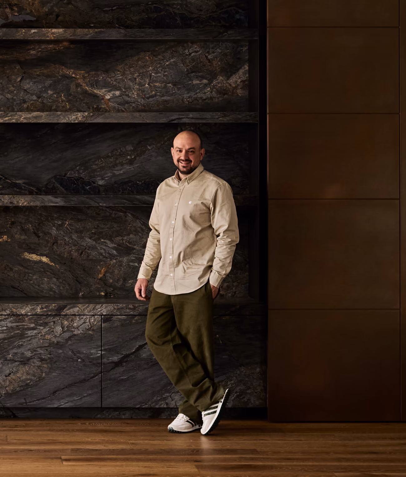 andrew loucaides, opus formed director, leaning against dark stone shelving beside bronze-toned panels