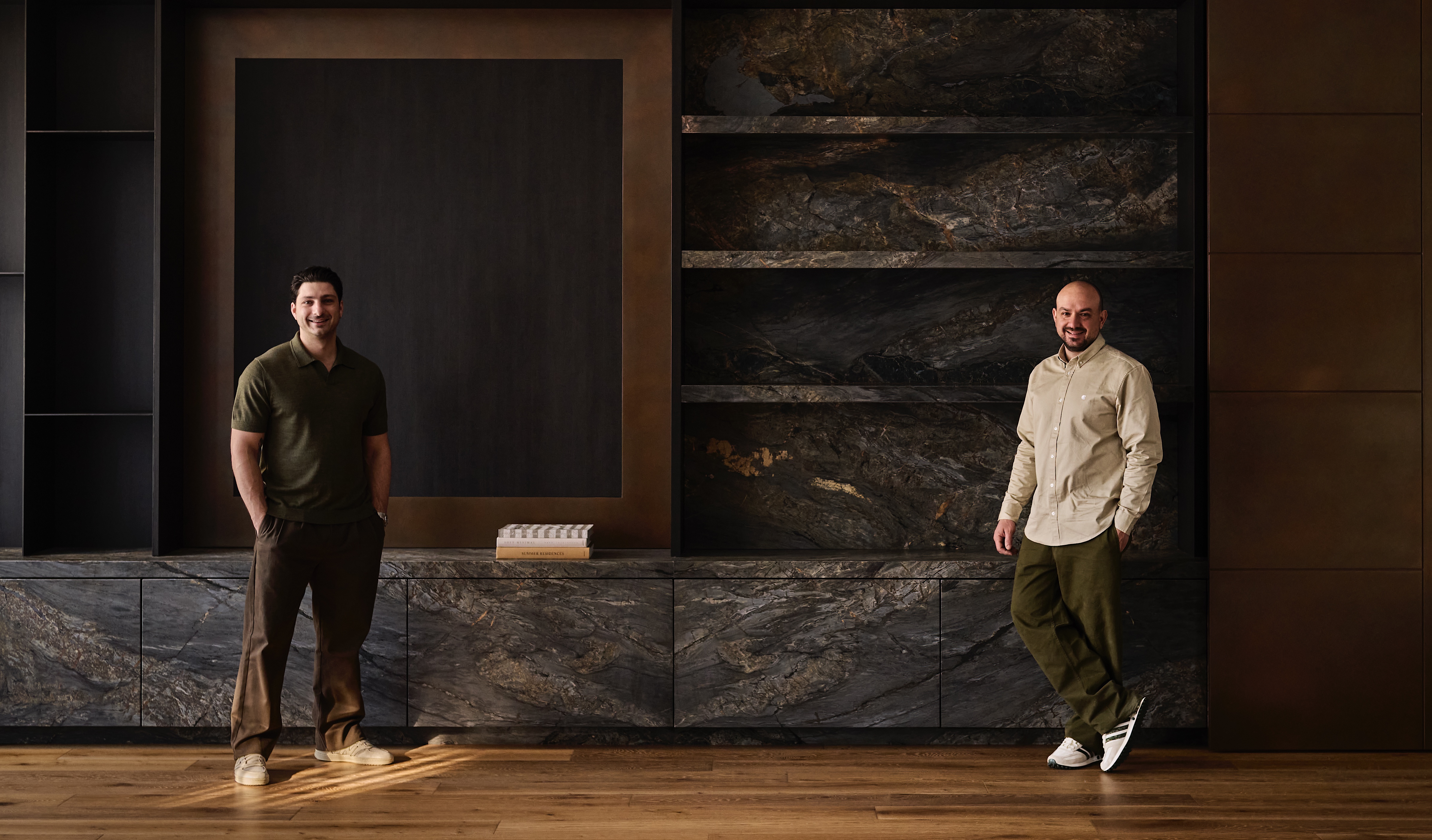 directors stevie hatz and andrew loucaides standing before dark stone shelving and bronze-toned panels