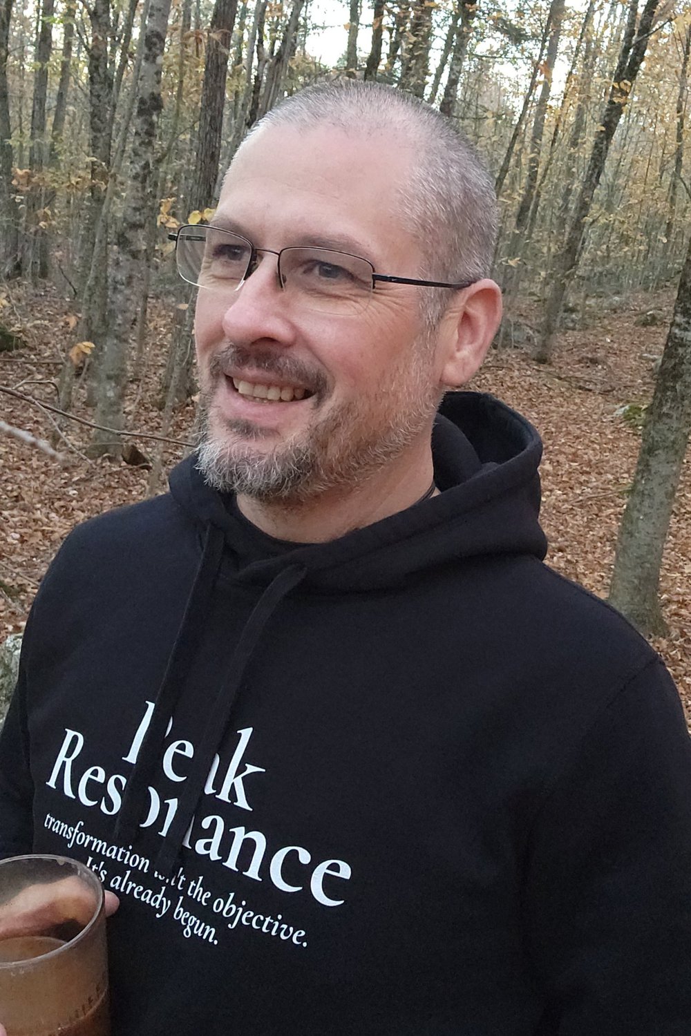 TJ, founder of Peak Resonance, smiling in the autumn woods in Maine, wearing a Peak Resonance hoodie and holding a coffee.