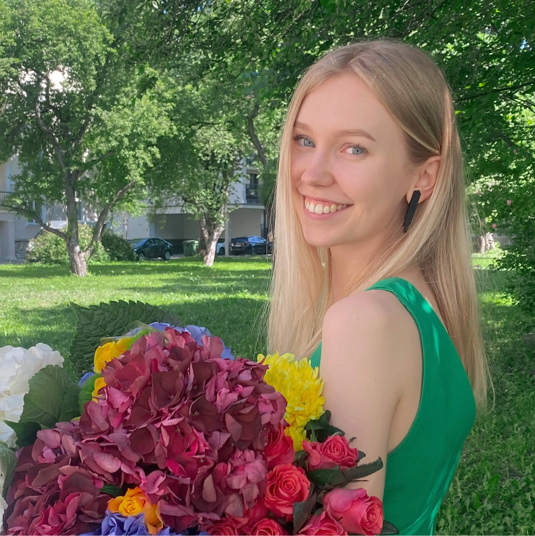 Woman with long blonde hair in a green dress holding a colourful bouquet of flowers outdoors on a sunny day.