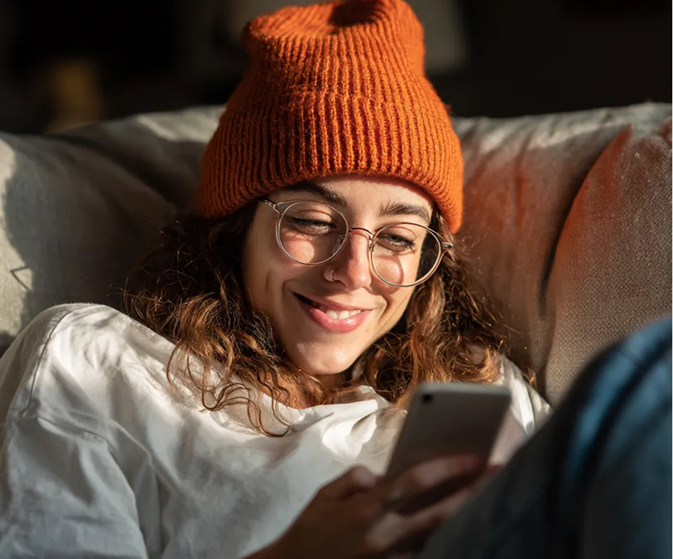 Junge Frau mit orangefarbener Strickmütze und Brille lächelt, während sie auf ihrem Handy schaut.