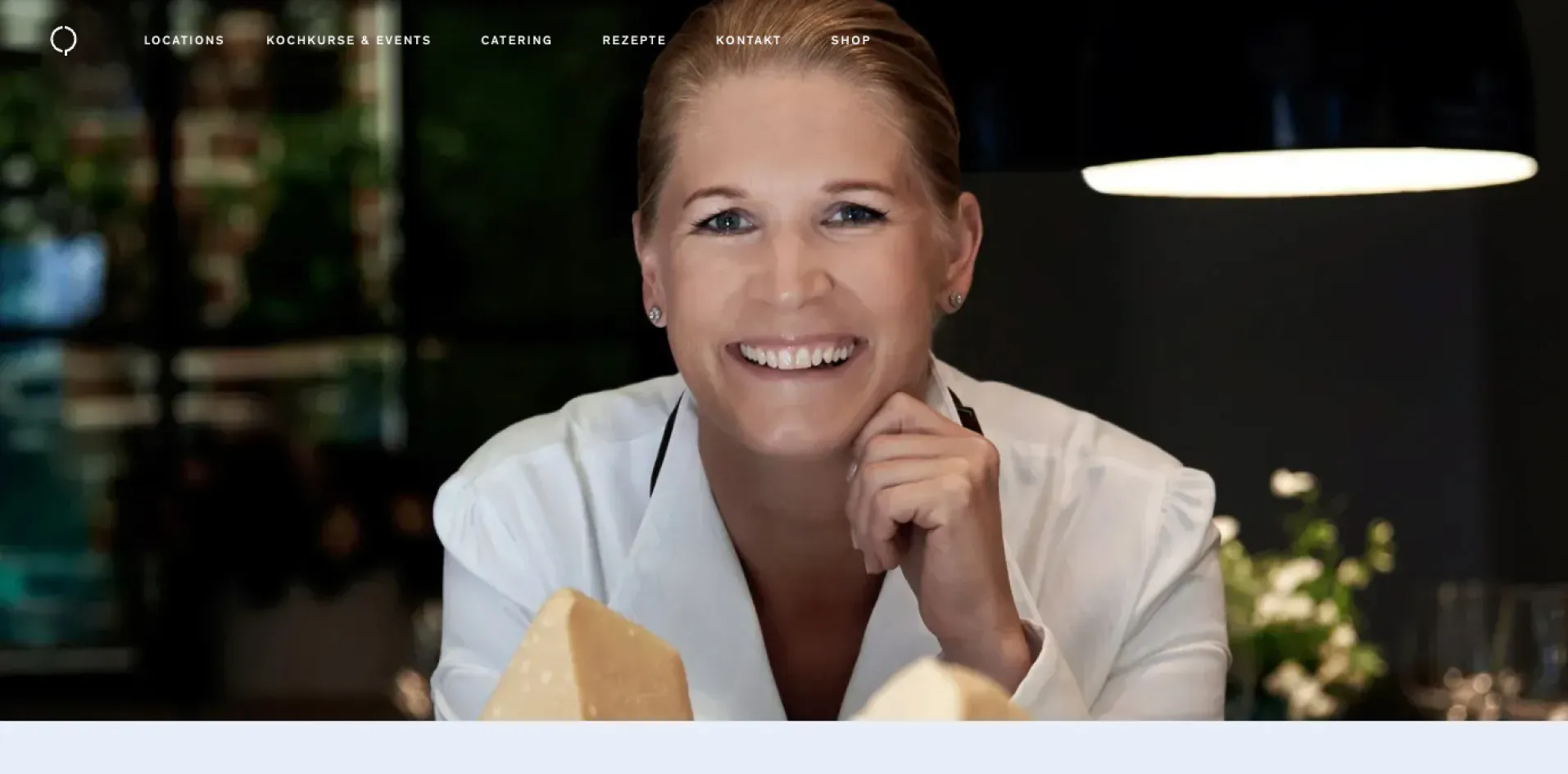 Lächelnde Frau in weißer Kochjacke mit zwei großen Käsestücken vor sich in einem gemütlich beleuchteten Raum.