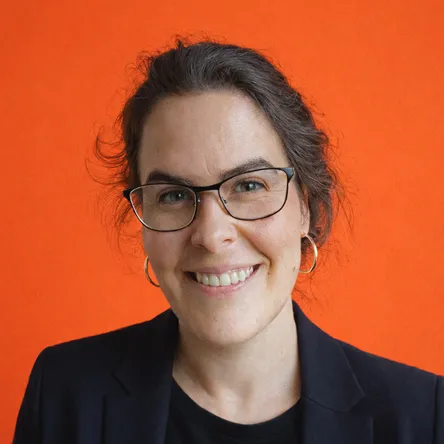 Eine lächelnde Frau mit Brille, dunklem Blazer und Ohrringen vor einem orangefarbenen Hintergrund.