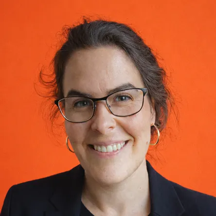 Porträt von mir mit einer lächelnden mit Brille, dunklen Haaren und dunklem Blazer vor orangefarbenem Hintergrund.