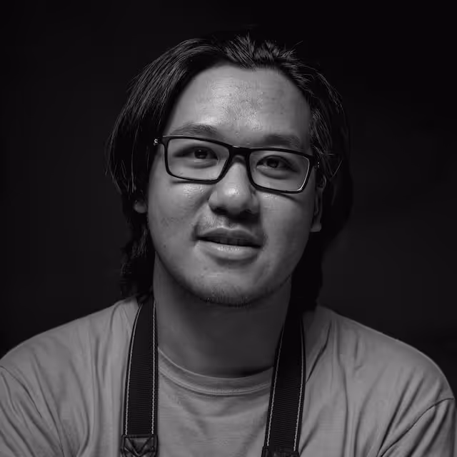 Black and white portrait of a man with medium-length hair, wearing glasses and a t-shirt, smiling slightly against a dark background.
