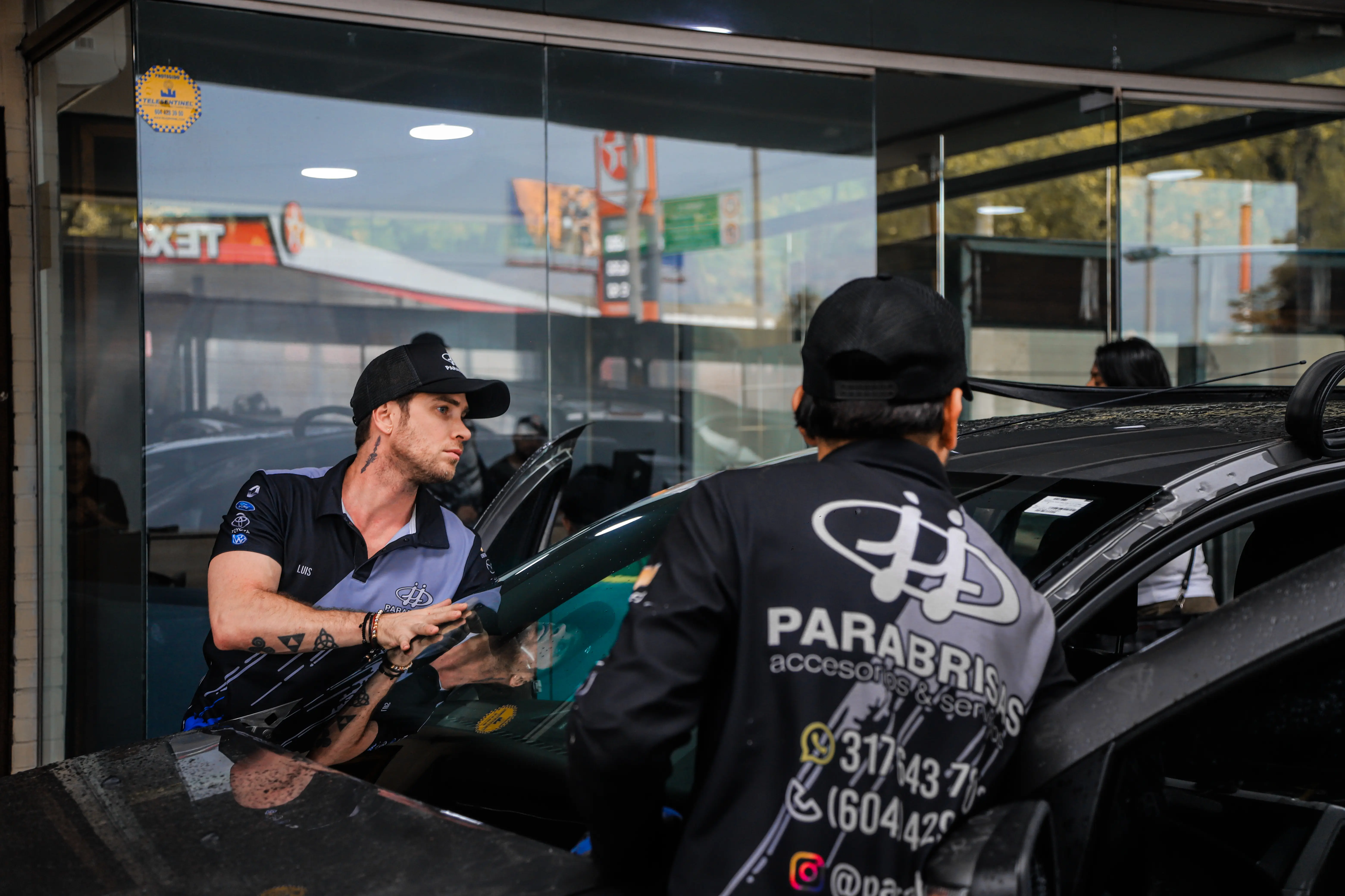Two men in black caps and Parabrisas uniforms working on a black car inside a glass-walled garage.