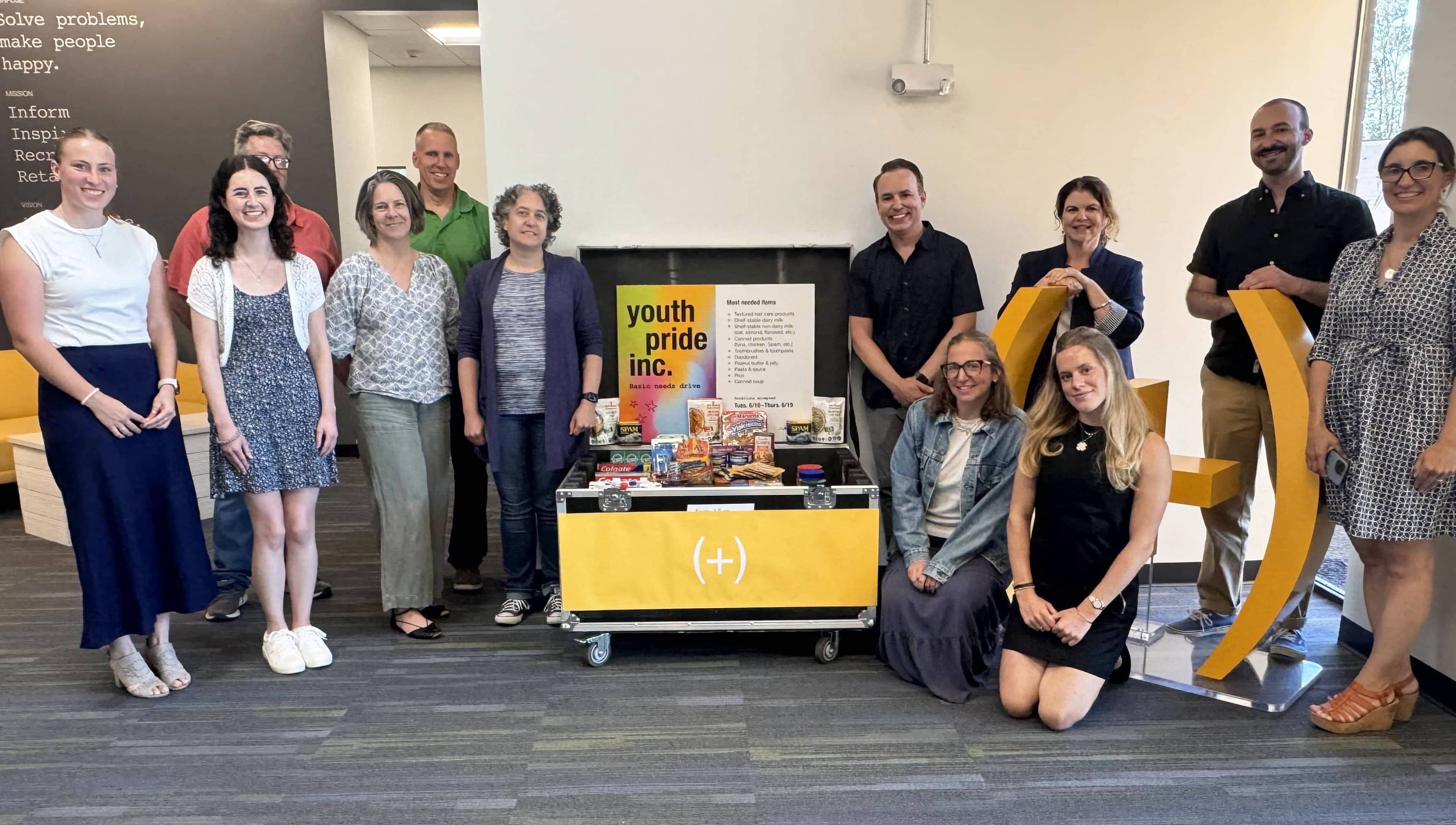 Team members at (add)ventures stand around a donation bin for Youth Pride Inc., collecting essential items in support of LGBTQ+ youth.
