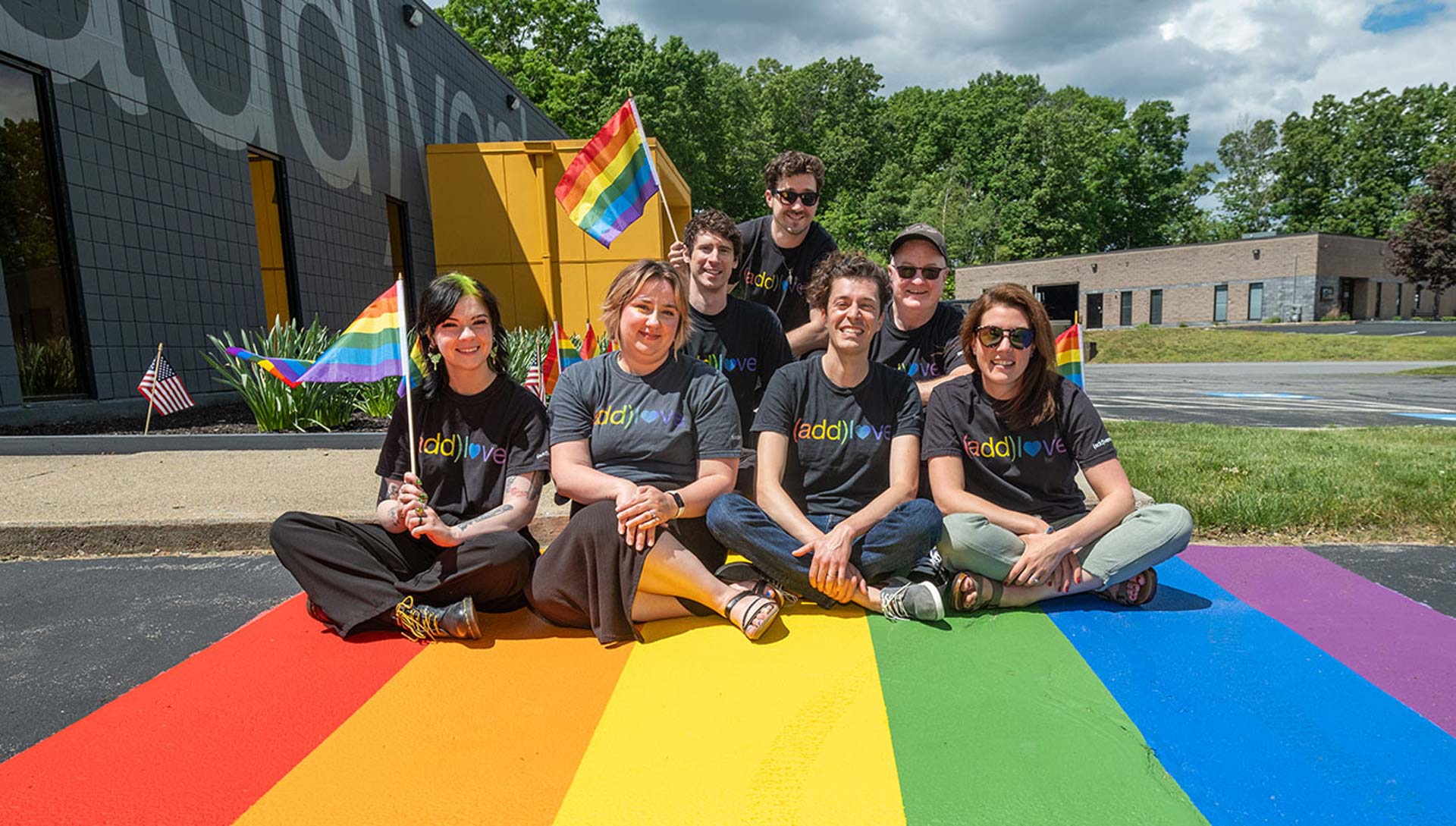Diverse group of people holding rainbow Pride flags, smiling and celebrating together outdoors under a clear sky.