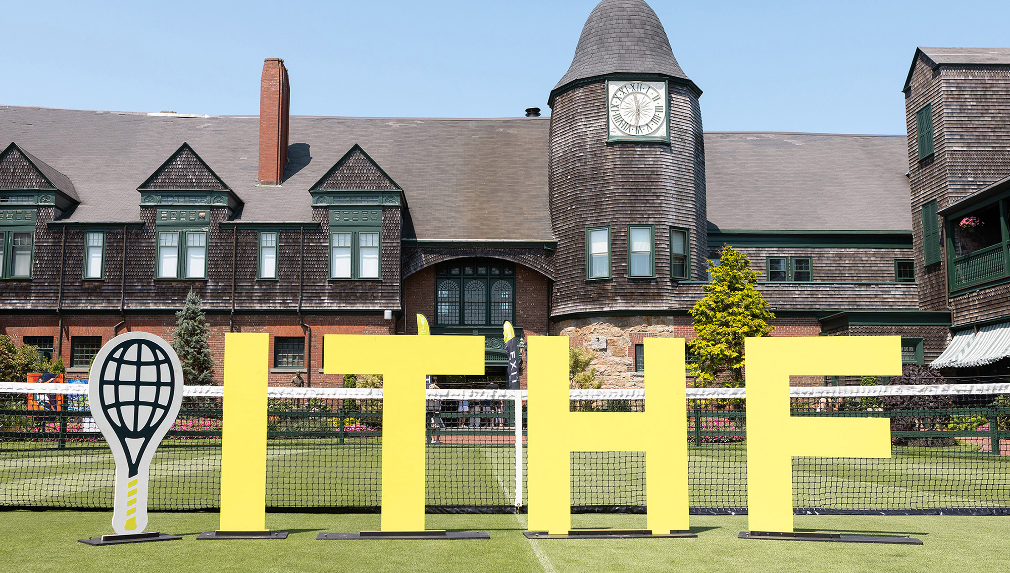  Large yellow letters spelling “ITHF” with a tennis racquet icon stand on a grass tennis court in front of the International Tennis Hall of Fame' historic shingle-style clubhouse.
