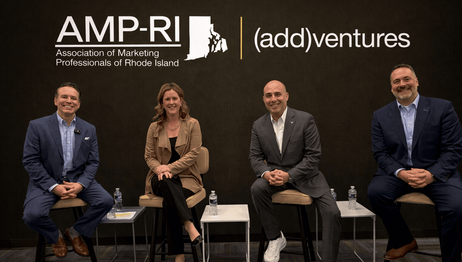 Four panelists, three men and one woman, dressed in business attire sit on stools facing the audience against a dark backdrop displaying the “AMP-RI – Association of Marketing Professionals of Rhode Island” and “(add)ventures” logos.