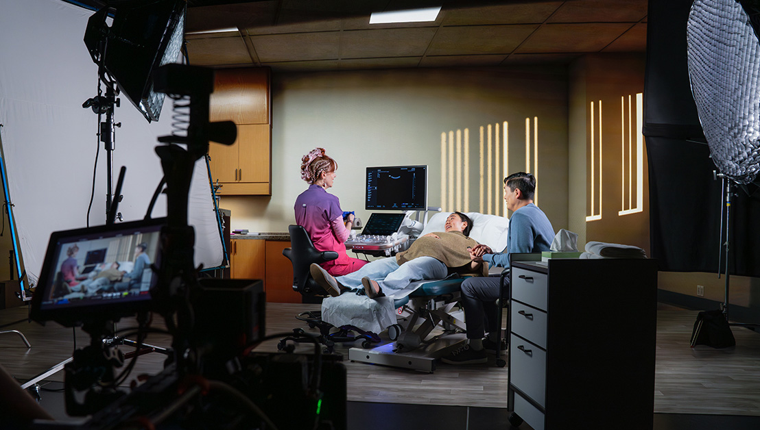 A medical exam scene being filmed: a clinician performs an ultrasound on a female patient lying on an exam table while a partner holds the patient’s hand, with professional lighting, cameras, and a monitor visible around the set.