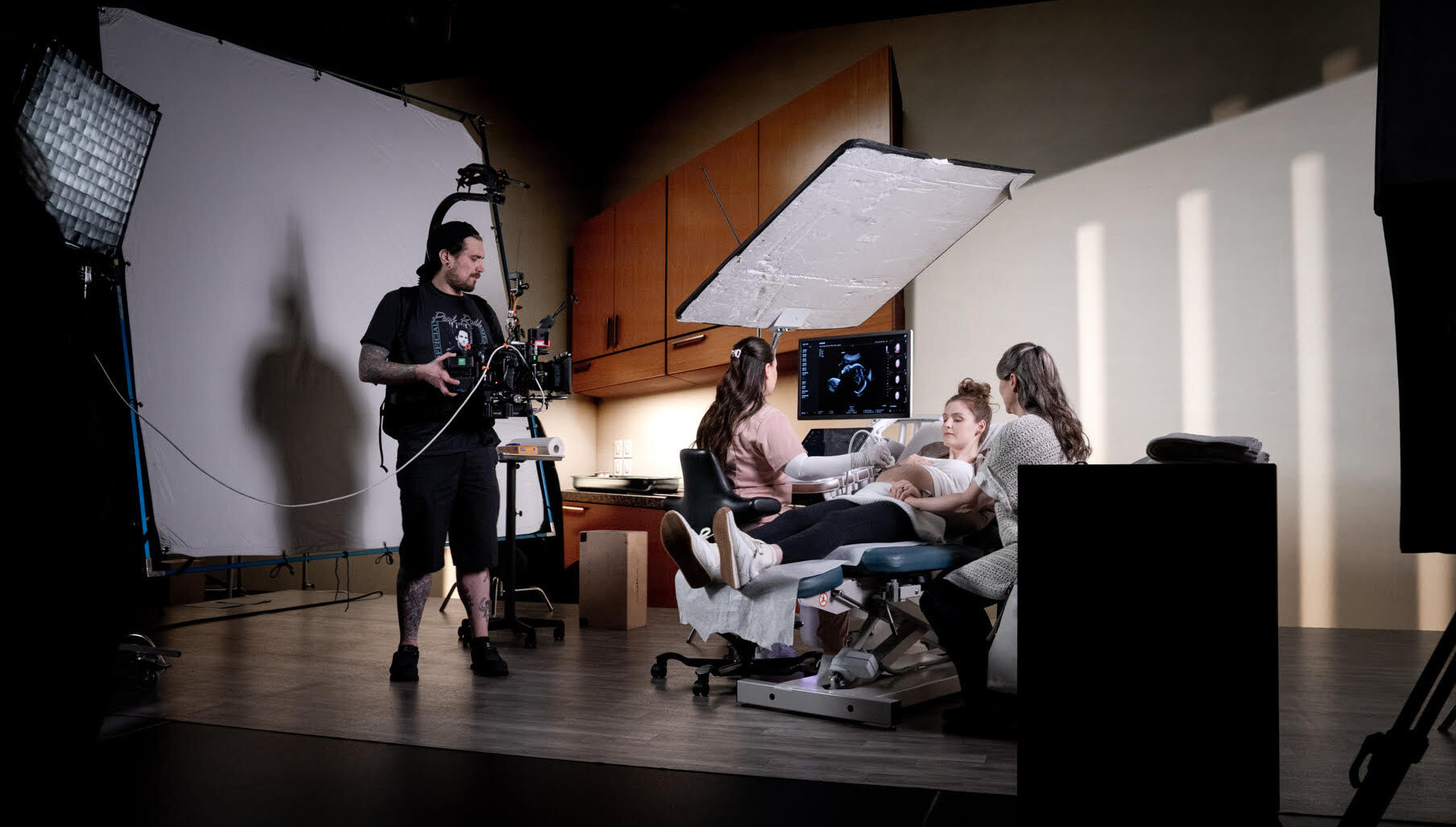 Camera operator films an ultrasound exam on a studio set, with a clinician scanning a patient and a companion beside them, against a staged medical room backdrop with cabinets and soft lighting and two blank screens for virtual projection.