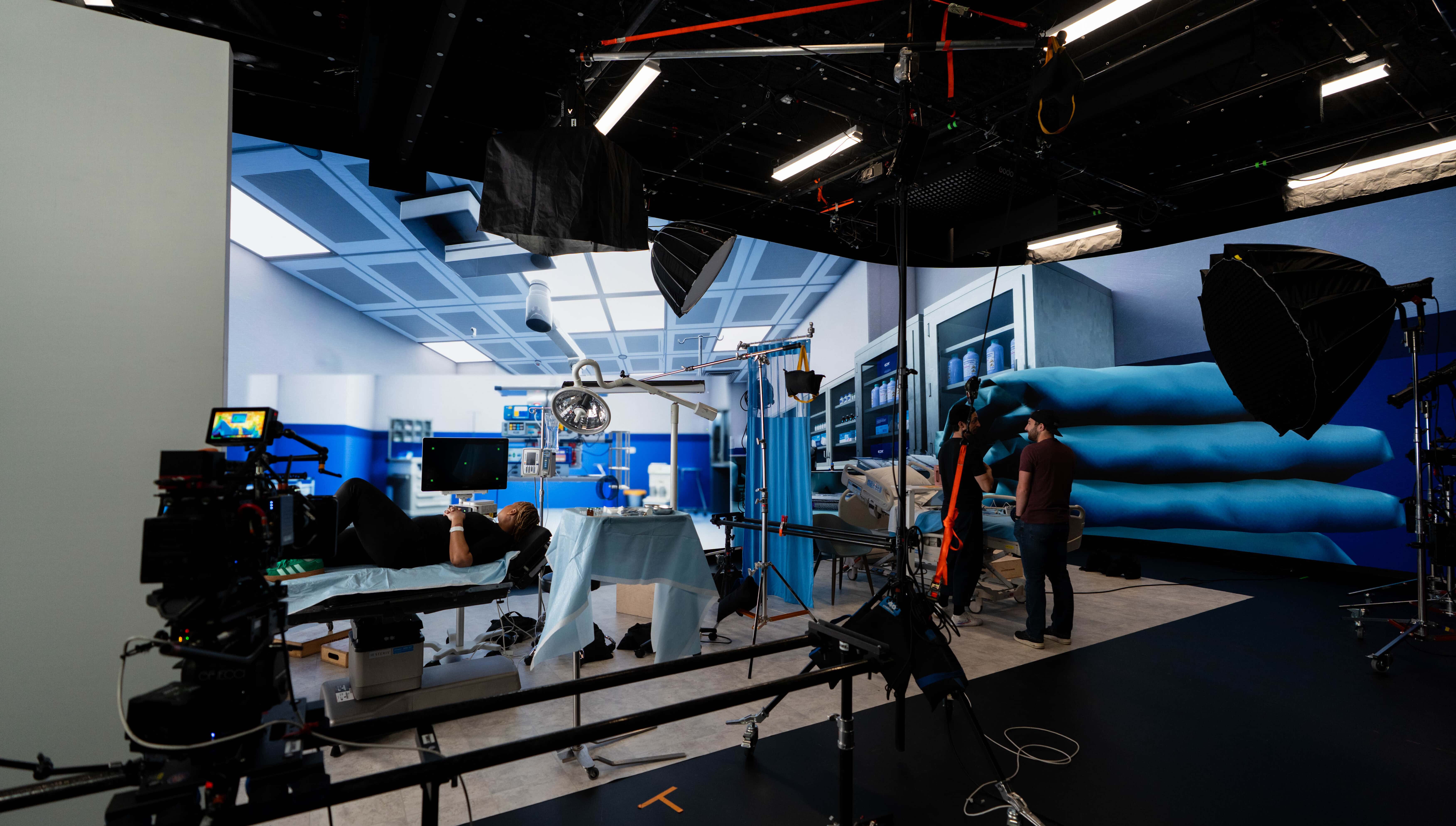 Wide view of a hospital set being filmed, with a patient on an exam table, production crew and equipment, and a large LED wall displaying a virtual medical room.