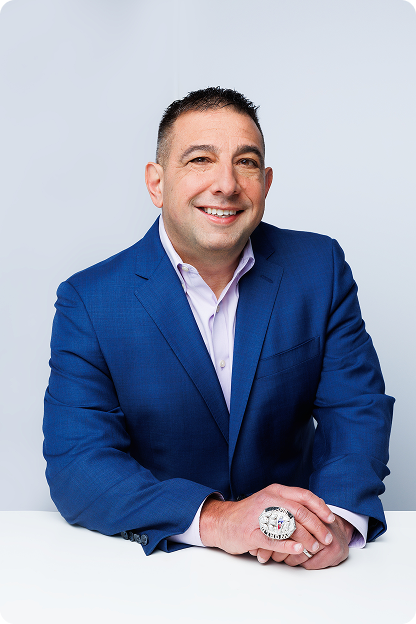Professional headshot of a smiling man in a blue suit jacket and light dress shirt, seated with hands folded wearing a Super Bowl LIII against a neutral gray background.