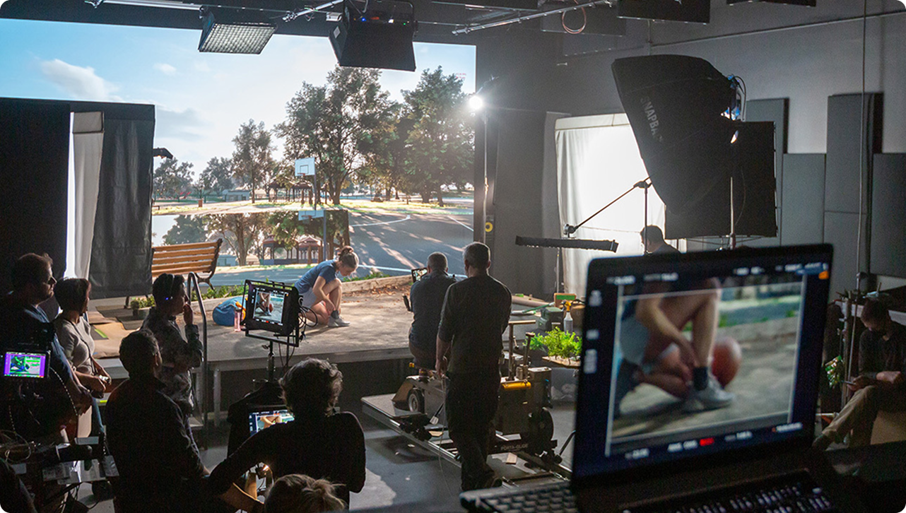 Film crew shooting a commercial on a soundstage using a large LED screen backdrop of a park scene; cameras, lighting rigs and monitors surround a person tying their shoe while crew members watch and adjust equipment.
