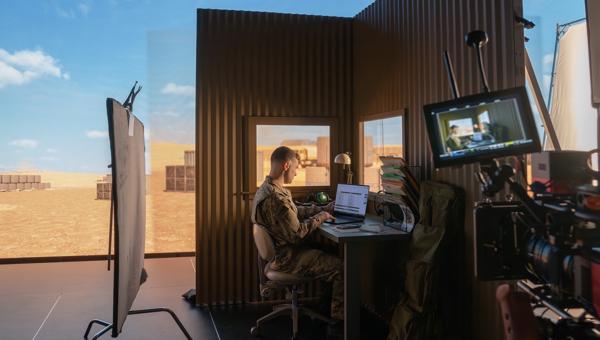 Behind-the-scenes video production of a military office set in a desert environment, with an actor in uniform working at a desk while a camera rig films the scene in the foreground.
