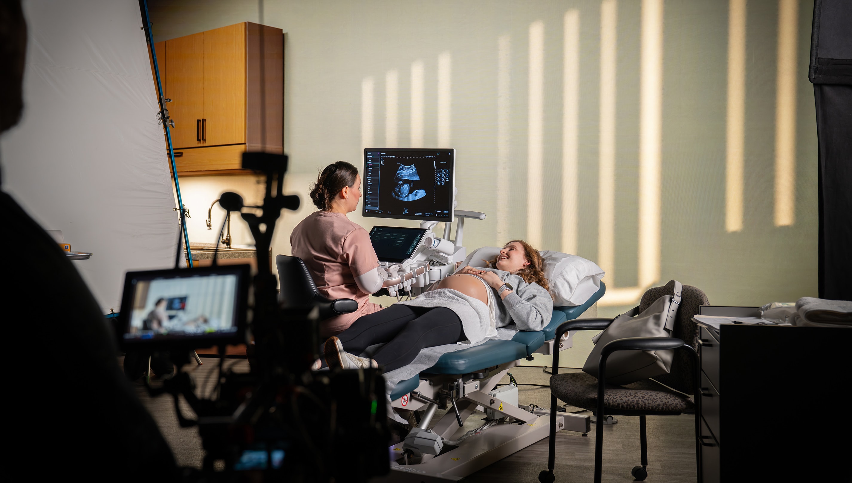 Behind-the-scenes video production of an ultrasound exam on a pregnant patient in a clinical setting, with a healthcare professional performing the scan while camera equipment films the scene from the foreground.