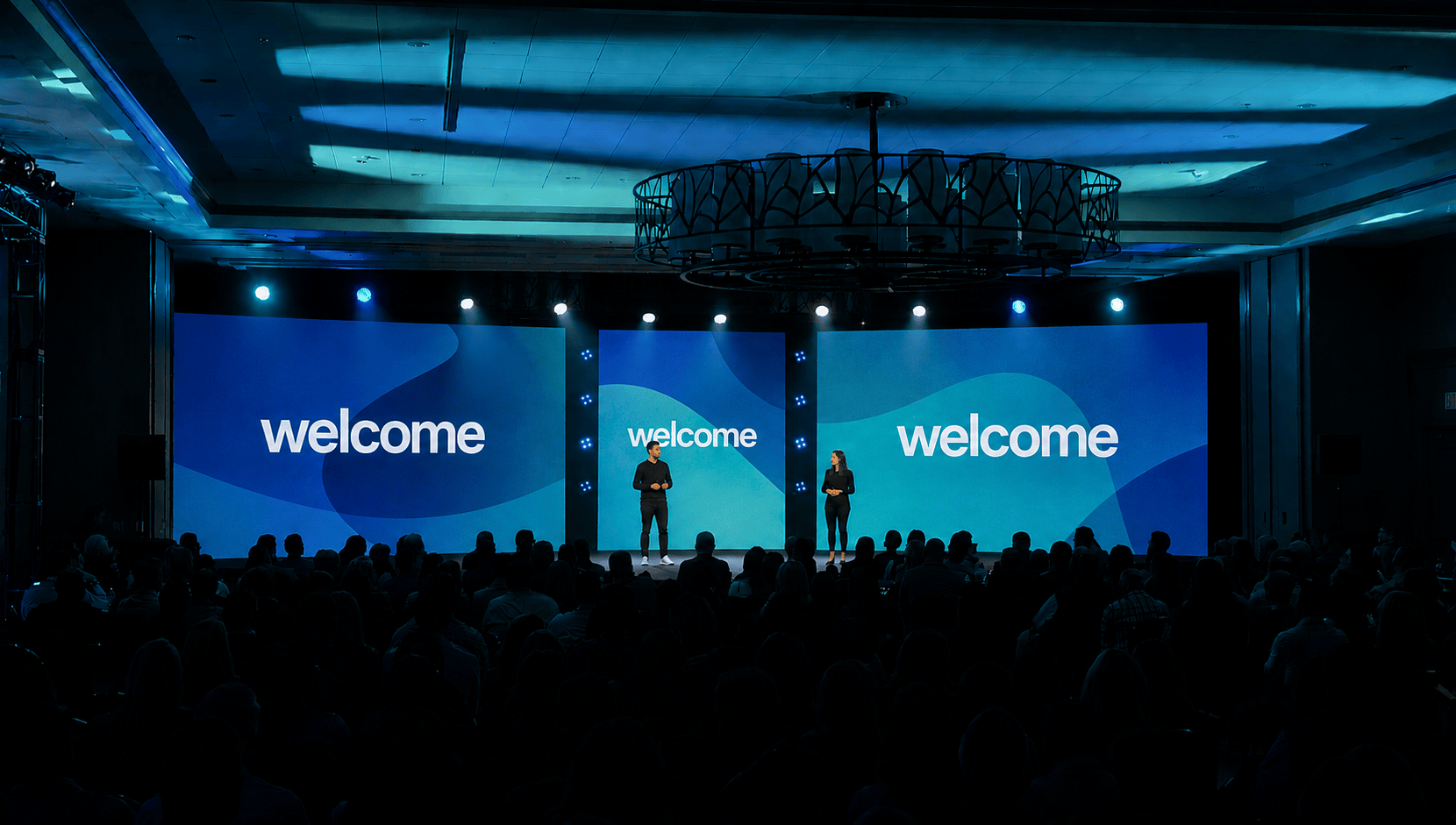 Wide conference stage with blue “welcome” screens, two presenters, and a seated audience in a dark ballroom.