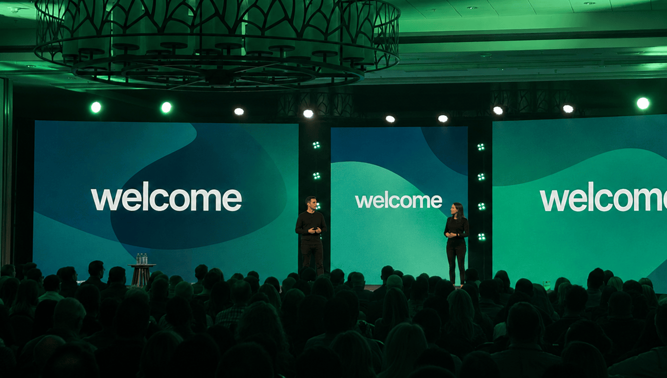 Event presenters stand before green “welcome” screens as an audience watches from a dimly lit conference room.