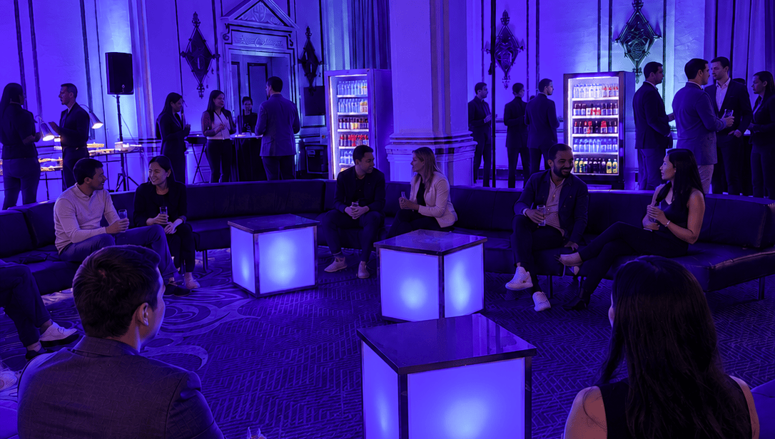 Attendees network in a purple-lit lounge with modern seating, glowing tables, and beverage displays.
