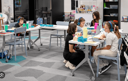 Students working independently on laptops in a modern classroom while a guide circulates; no teacher at the front of the room.
