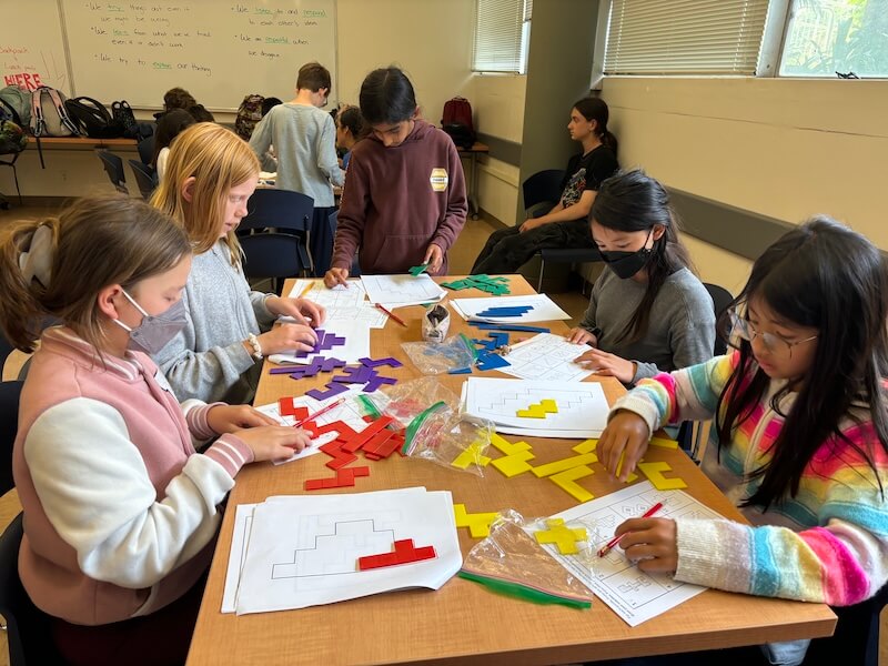A diverse group of elementary school children smiling and solving interactive math puzzle games together with a high school student mentor at a local library table.