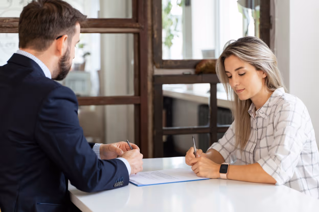 A man in a suit and a woman with long blonde hair sit across from each other at a table, reviewing and signing documents in an office setting.