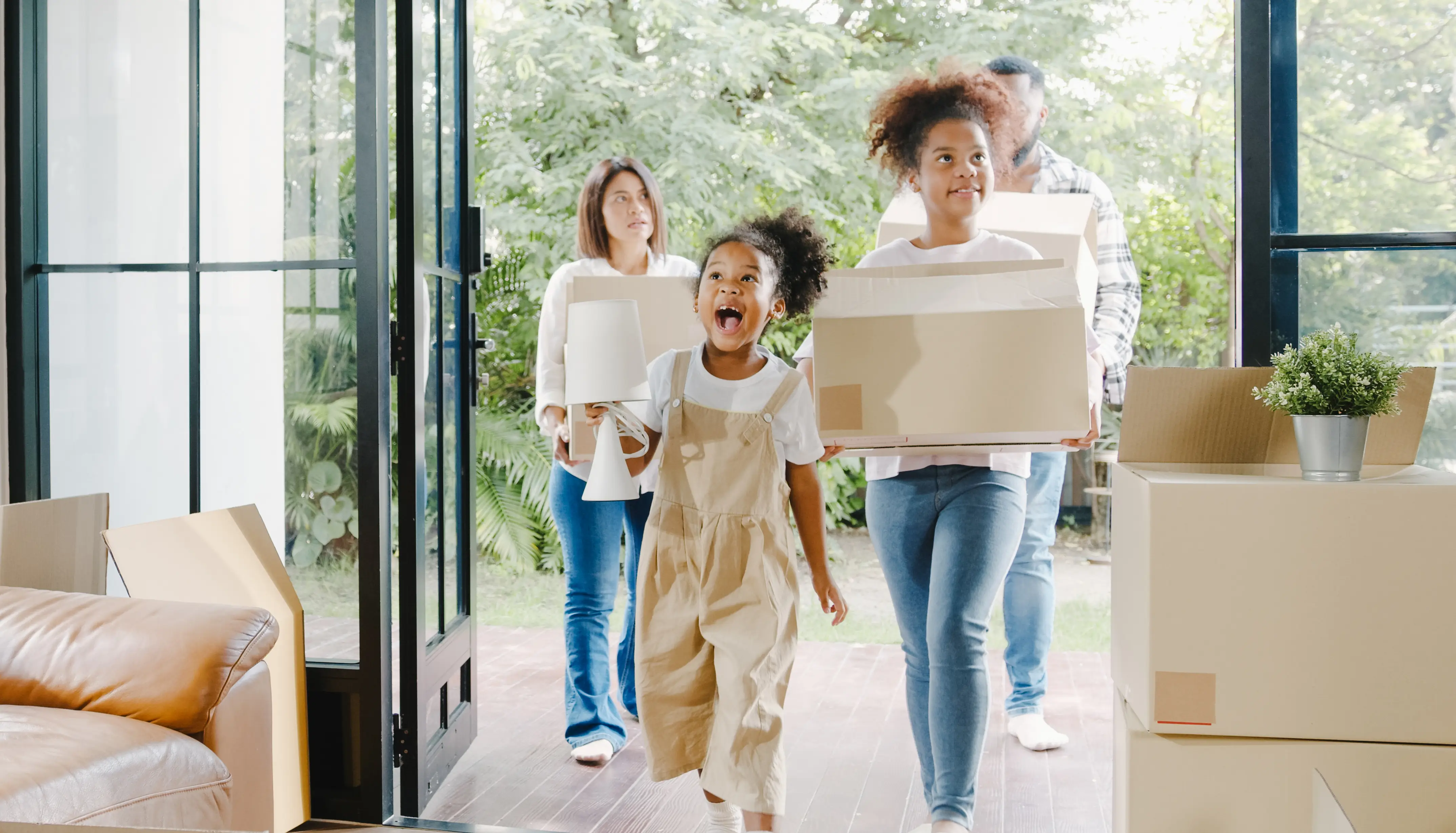 A happy family carrying boxes and belongings walks into a bright home, with a smiling young girl leading the way. Green trees are visible outside through the open doorway.