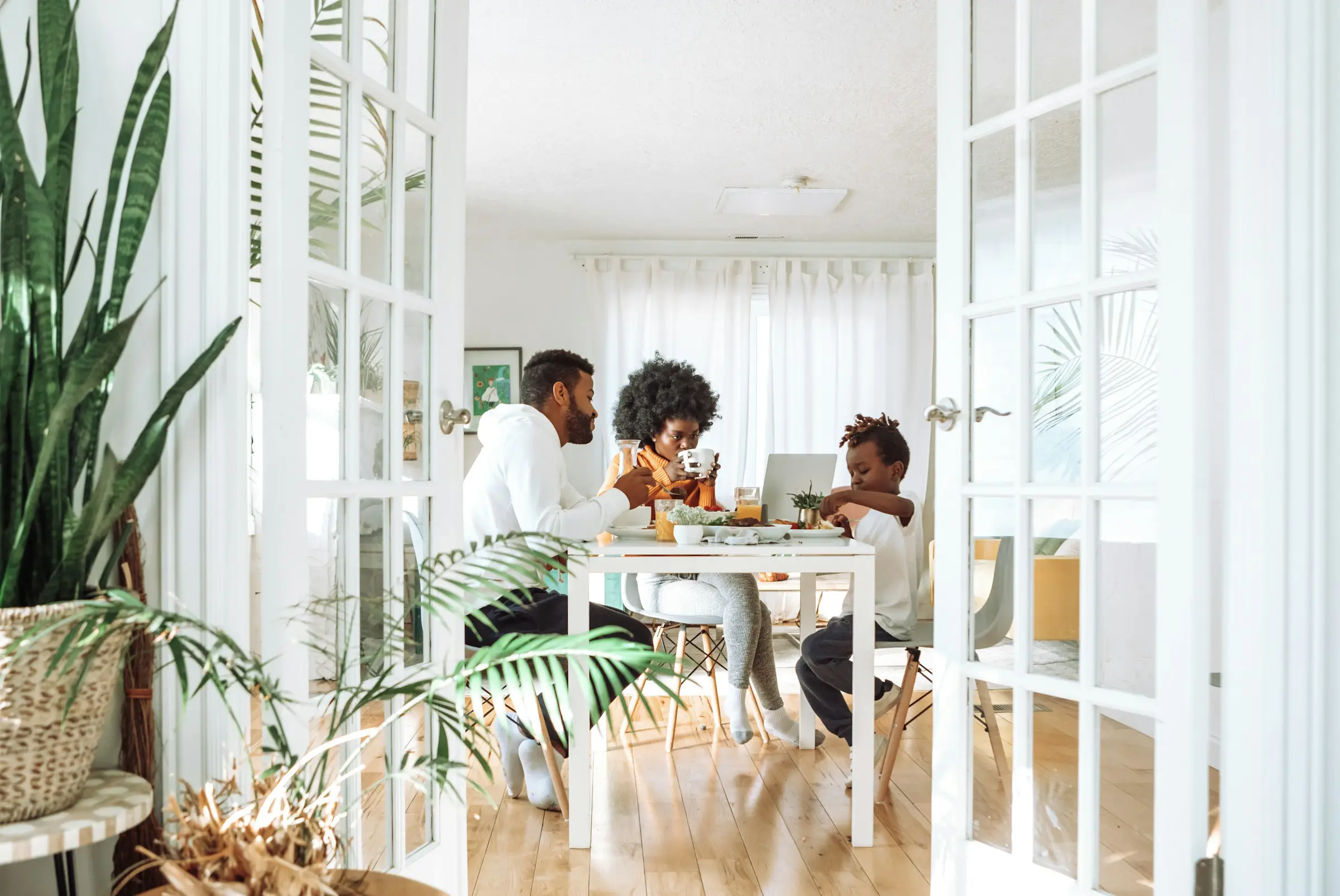 A family of three sits at a dining table in a bright, sunlit room with large windows and indoor plants, sharing a meal together.