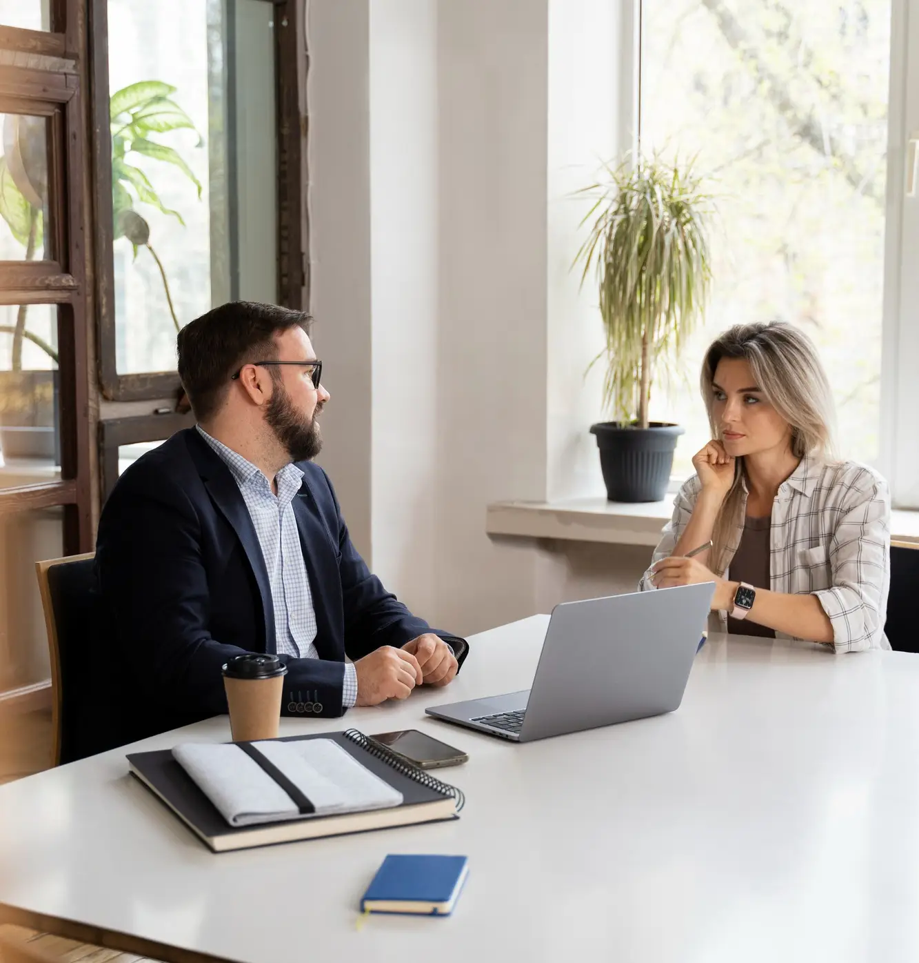 A man and a woman sit at a white desk with a laptop, notebook, and coffee cup. They are having a serious conversation in a bright office with large windows and potted plants.